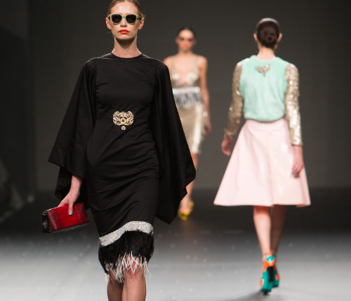 Model walking on a fashion show runway wearing a black outfit with a belt.