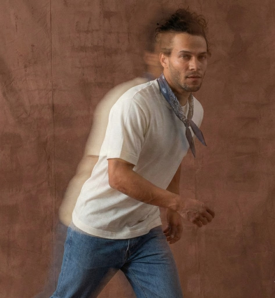 Man in a white t-shirt and blue jeans with a bandana, running against a textured brown wall.